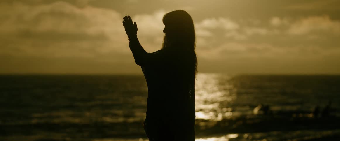 Silhouetted woman standing on the shore raises her hands prayfully to the sky while teams race boats in the background on a cloudy day