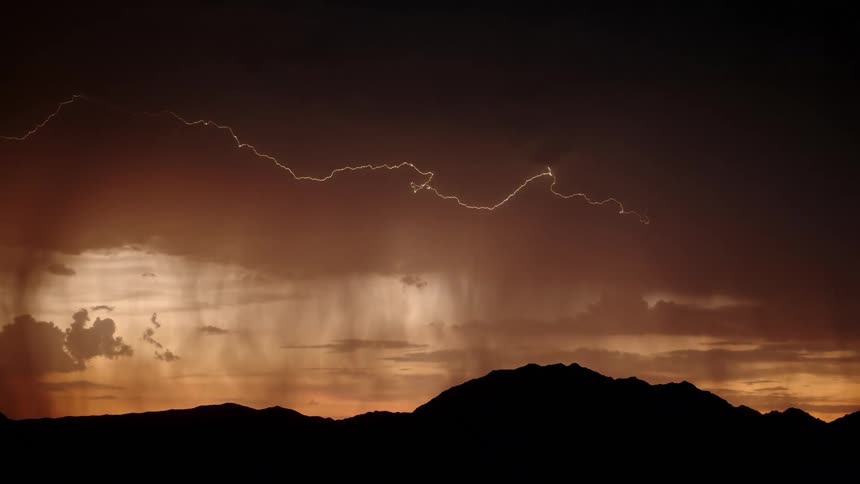 Forks of lightning dramatically illuminate a stormy sunset sky as sheets of rain fall over a silhouetted mountain range