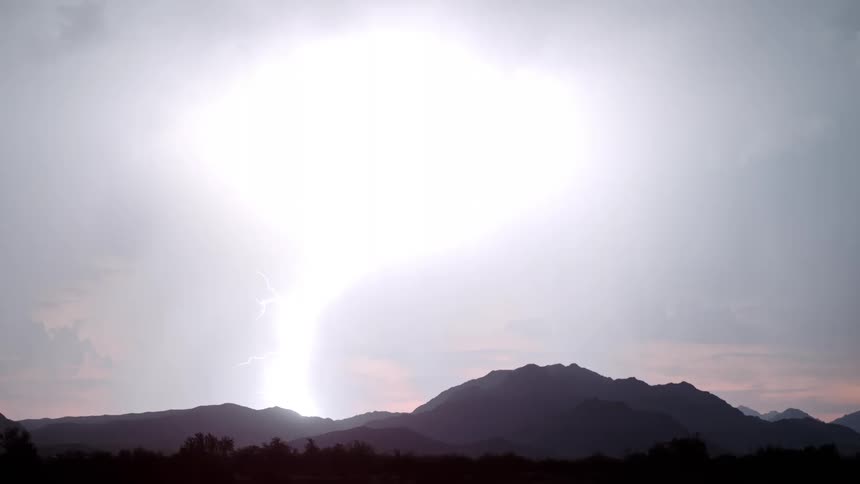 A bright bolt of lightning strikes across a dark night sky above a silhouette of a mountain range during a thunderstorm while orange light glows along the horizon and rain falls in the distance