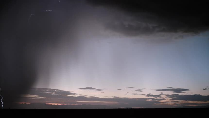 A dark thunderstorm looms over a flat horizon at twilight while bolts of lightning flash through a heavy curtain of falling rain against a fading purple and blue sky