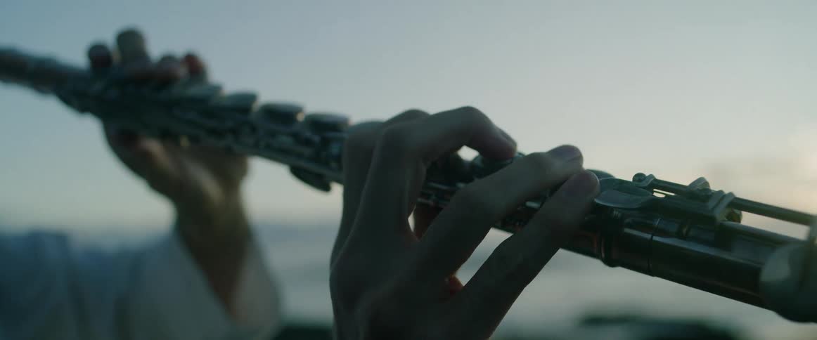 A woman plays a silver flute outdoors against a coastal sunset with the camera focusing on her fingers moving over the keys as the sun glows over the ocean and rocky shoreline in the background