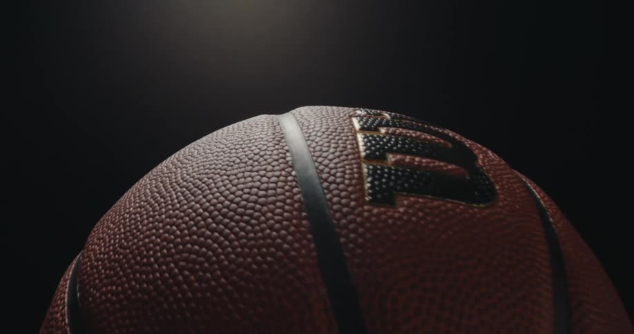 Extremely close view of the pebbled orange surface and black ribs of a basketball rotating slowly under a bright spotlight against a dark background