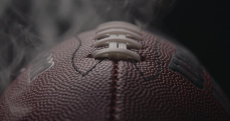 An extreme macro shot captures a brown american football with white laces as thin wisps of smoke rise from its pebbled leather surface against a dark moody background