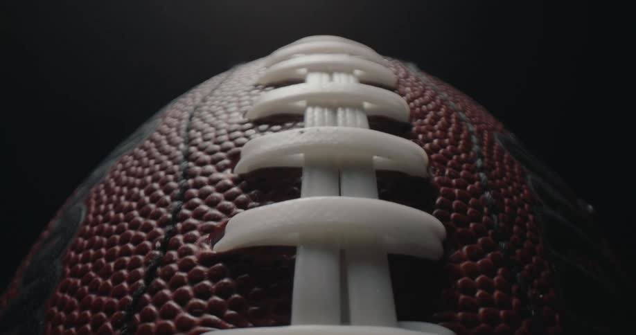 A close view of the white laces and textured brown surface of an american football under a single light source against a dark background