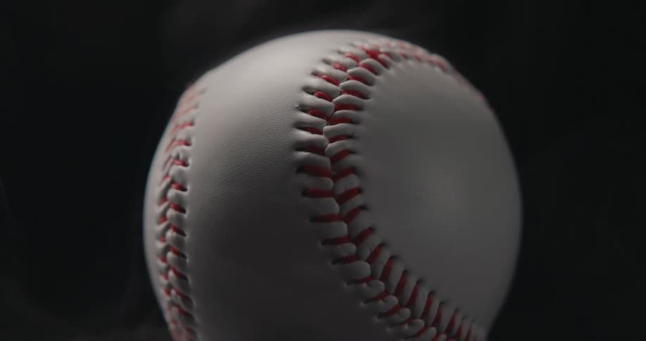 A baseball rotates slowly against a dark moody background while wisps of smoke curl around its white leather surface and red stitching in a tight closeup