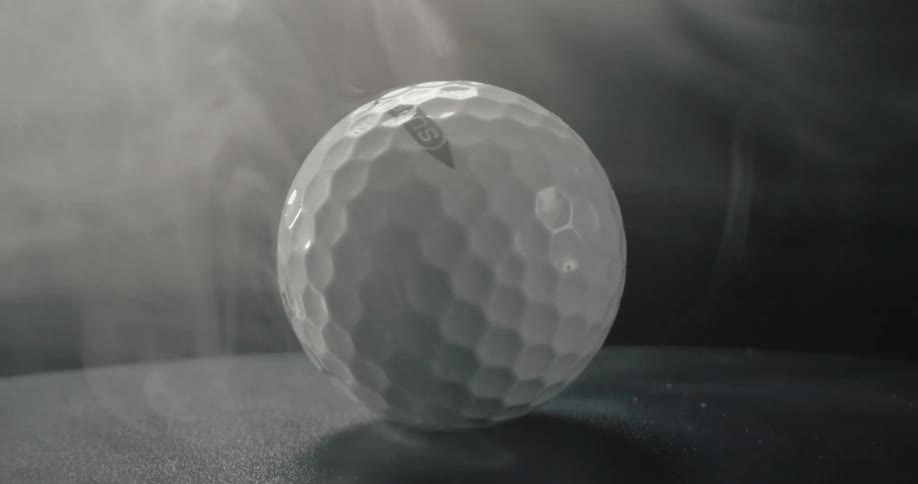 A white golf ball rotates slowly on a dark flat surface while thick white smoke drifts across the frame under a bright overhead spotlight against a black background