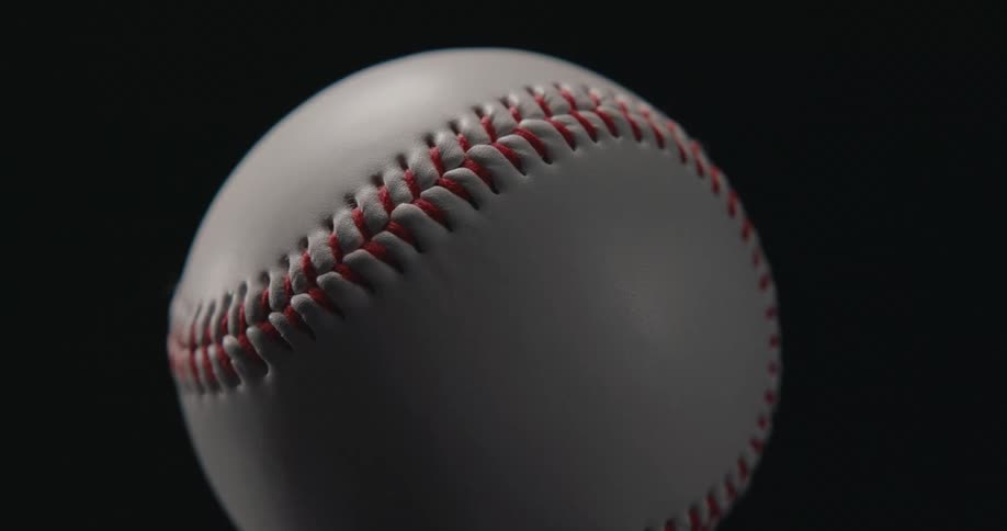 A macro shot shows a white baseball with red stitching rotating slowly against a solid black background highlighting the texture of the leather and the detail of the seams as it spins in a dark studio setting