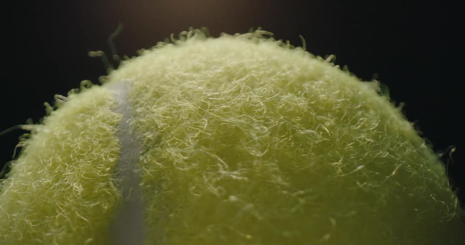 An extreme closeup video shows a yellow tennis ball slowly rotating against a dark background the camera focuses on the detailed texture of the fuzzy neon fibers and the white seam as it moves