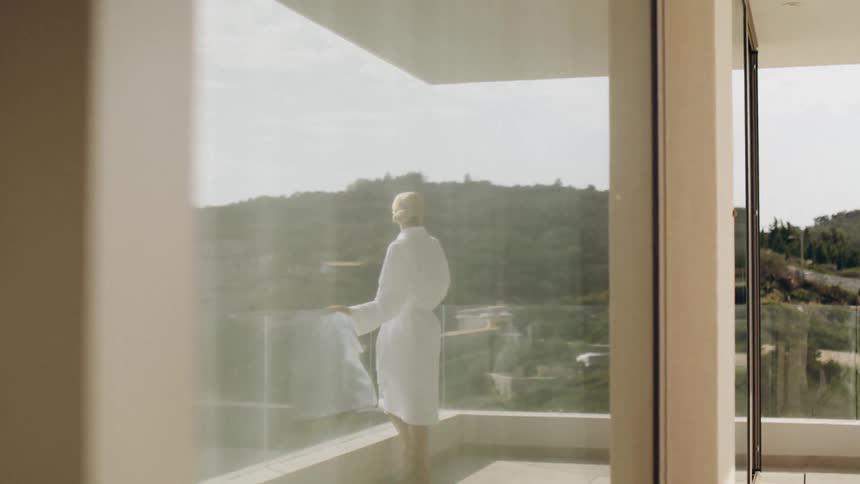 A view through a glass window captures a woman with short blonde hair wearing a white bathrobe standing on a balcony looking out at a green hill in the distance