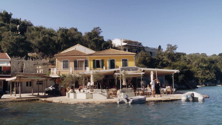 A sunlit waterfront caf and yellow villa sit along a calm shoreline as people relax beside the water and small boats