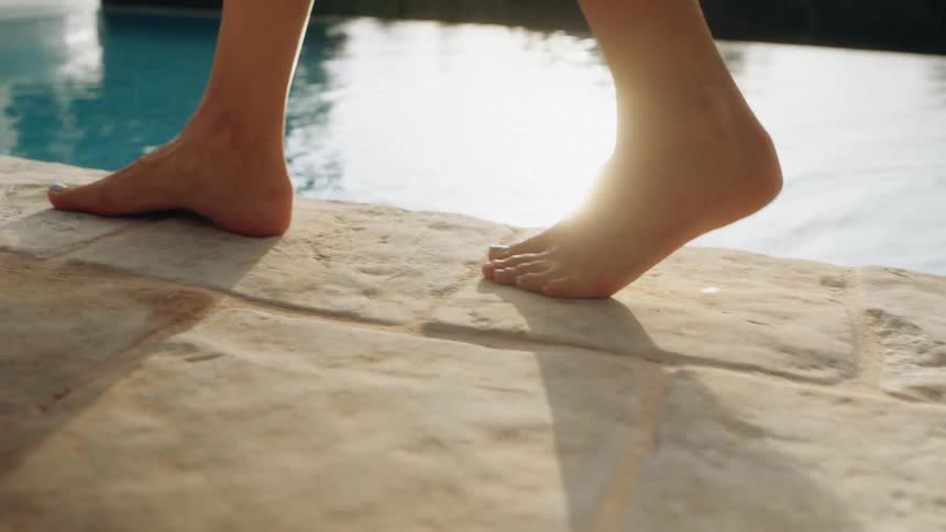 Female person walking next to a swimming pool and dipping their feet on the water outdoors during dusk or dawn