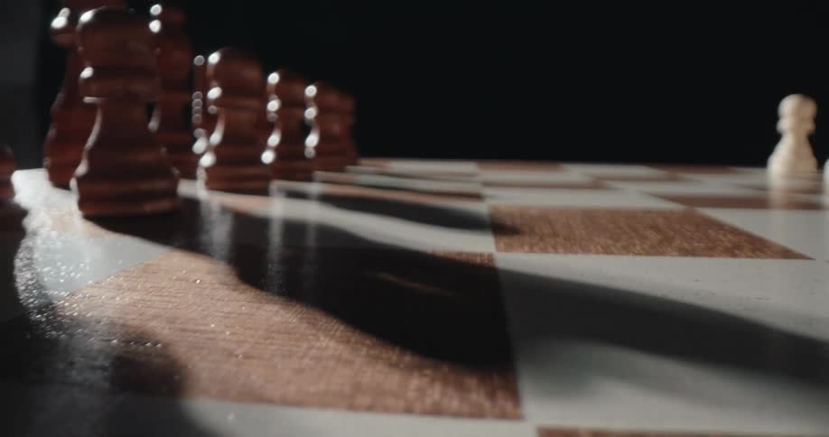A lowangle macro shot shows wooden chess pawns lined up on a board as long shadows sweep across the squares while white pieces are visible in the distant background against a dark void