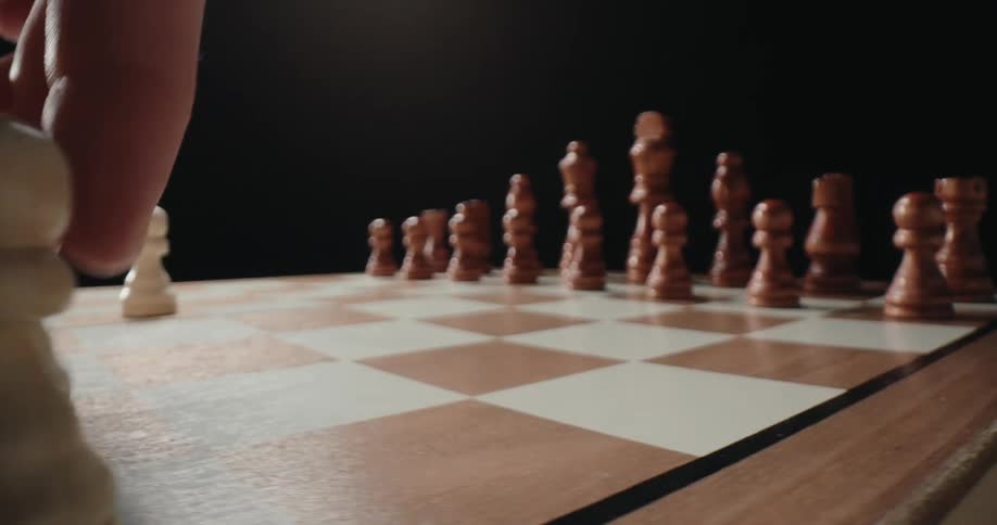 A hand reaches into the frame and moves a white pawn forward on a wooden chessboard to begin a game while the brown pieces remain lined up in the background against a dark setting