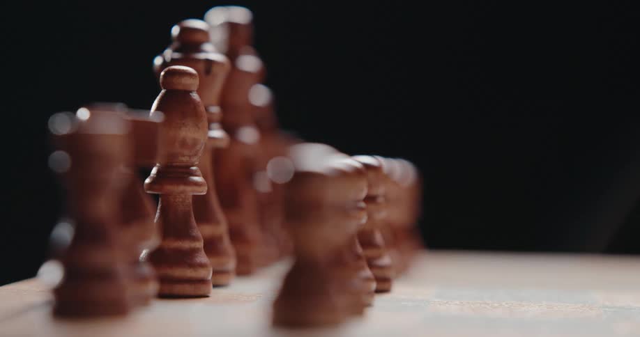 A macro lens shifts focus along a row of wooden chess pieces standing on a board against a dark background starting on a bishop and moving toward the pawns
