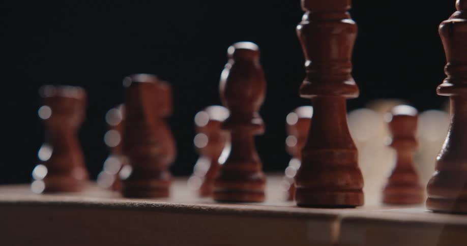 Wooden chess pieces stand on a wooden chessboard in a dark area