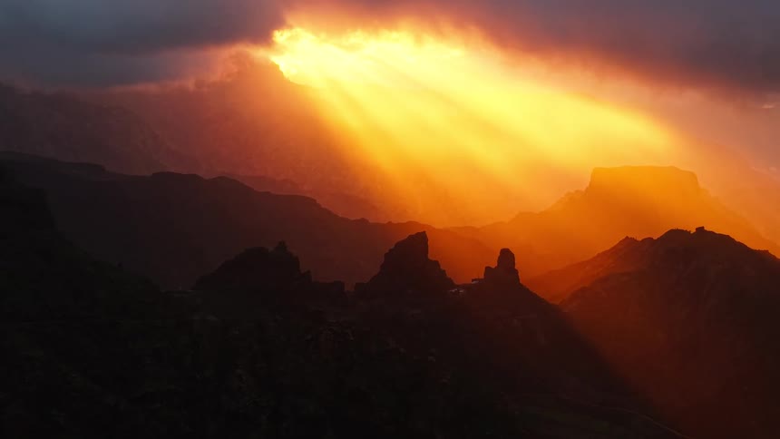 Bright orange sunbeams break through dark clouds to illuminate a jagged mountain range casting deep shadows over the valleys and creating a highcontrast landscape as the light shifts across the silhouetted peaks and rocky canyon walls