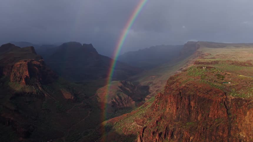 Aerial view of a rocky canyon with a bright rainbow spanning across a stormy dramatic sky