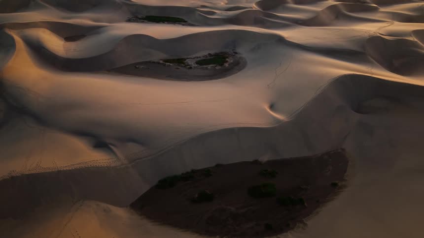 An aerial camera slowly pans up over vast golden sand dunes at sunset revealing the expansive desert landscape meeting the edge of the ocean