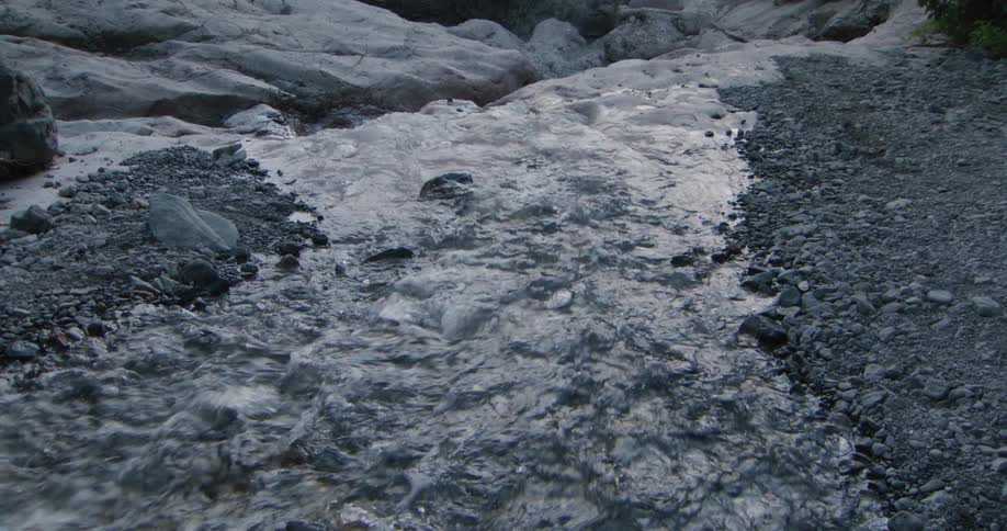 A clear shallow stream rushes quickly through a gray rocky canyon its water tumbling over smooth pebbles along the creek bed