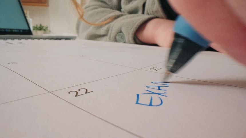 A person uses a blue marker to write the word exam on the 23rd day of a large desk calendarthe camera angle provides a closeup perspective of the hand moving across the paper while a laptop sits open in the background