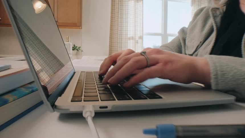 A young woman with red hair sits at a kitchen counter and types on a laptop computer the camera zooms in closely to show her fingers pressing the keys on the keyboard