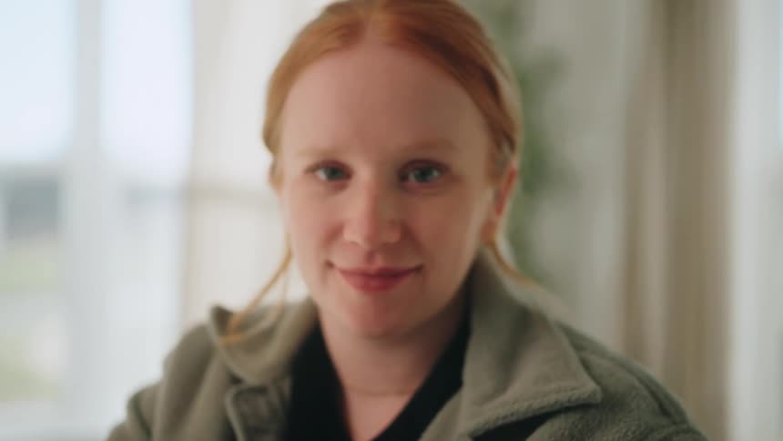 A young woman with red hair looks down before raising her head to look directly at the camera with a gentle smile as the image shifts from blurry to sharp focus in a bright indoor room