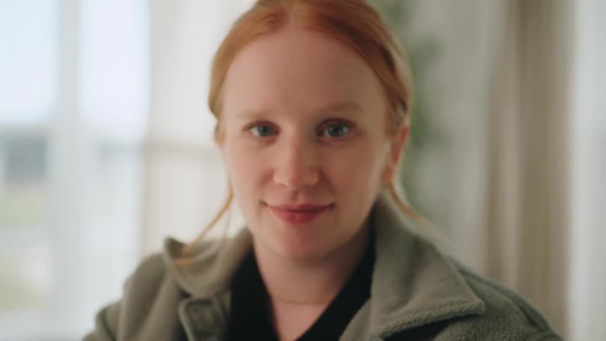 A young woman with red hair wearing a green jacket sits with a smile on her face