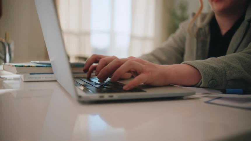 A woman wearing a grey sweater sits at a white desk and types on a silver laptop keyboard