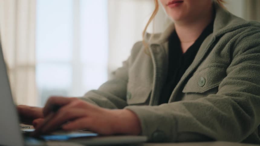 A redheaded woman wearing a green fleece jacket sits at a white desk and types on a laptop