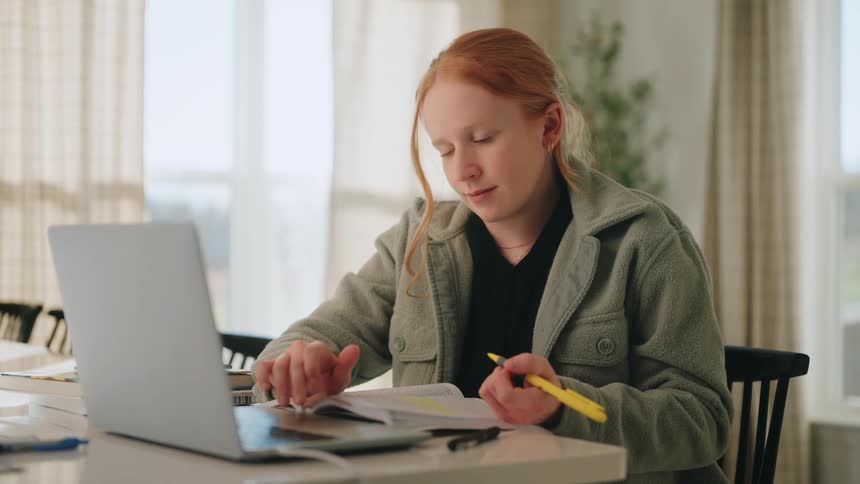 A young woman with red hair sits at a table using a laptop while highlighting text in an open textbook with a yellow marker