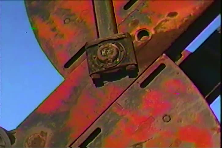A weathered red metal agricultural blade with bolts and scratches rotates against a clear blue sky its rust and chipped paint giving it a gritty vintage industrial feel