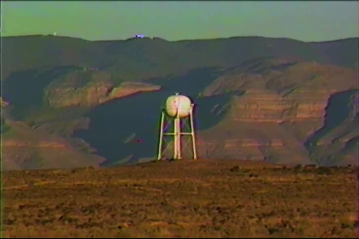 A white water tower stands on a hill in a vast desert landscape with large mountains in the background