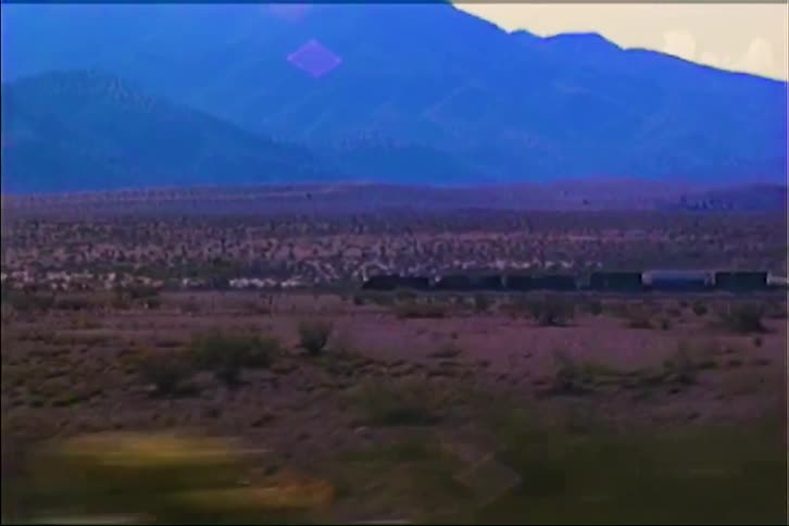 The video shows a long freight train moving across a wide desert valley during the late evening blue mountains stand in the distance under a hazy sky while the foreground is filled with dry brush and lowlying vegetation