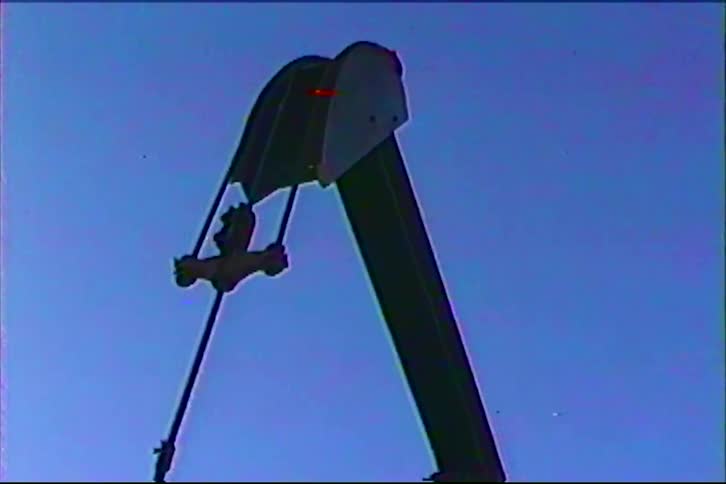 The video shows the silhouette of an industrial oil pump jack moving rhythmically against a clear blue sky the footage has a vintage or retro aesthetic characterized by visual artifacts and color distortion similar to an old vhs tape