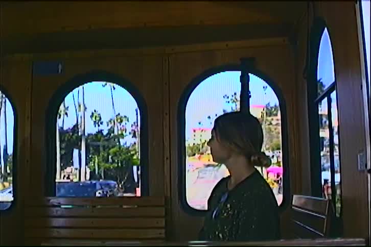 A woman sits quietly inside a wooden trolley car turning her head to look out through the arched windows at palm lined streets bathed in bright daylight evoking a nostalgic leisurely urban ride