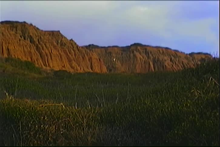 Steep rugged brown cliffs rise above dense green shrubs under a pale sky