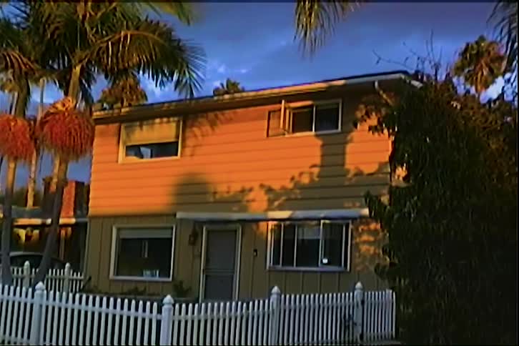A twostory tan suburban house stands behind a white picket fence and palm trees during sunset as shadows from nearby foliage slowly crawl across the front facade