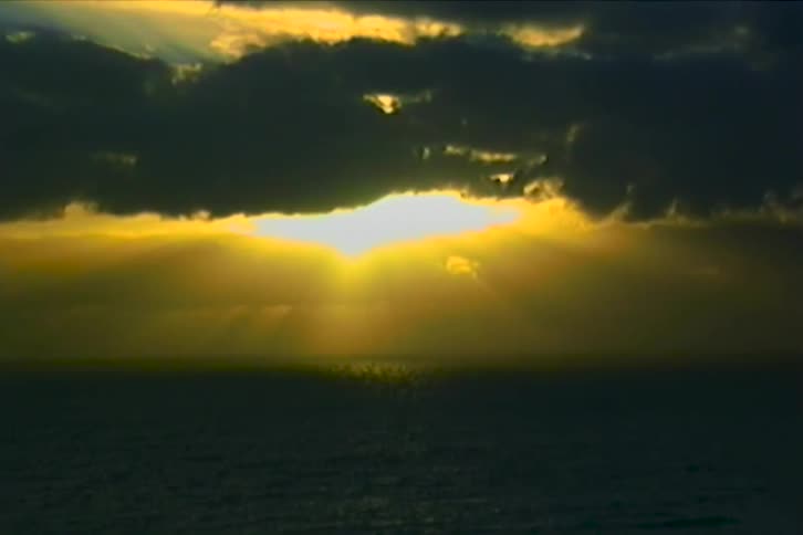 Dark clouds partially obscure the glowing sun as it sets over a rippling ocean