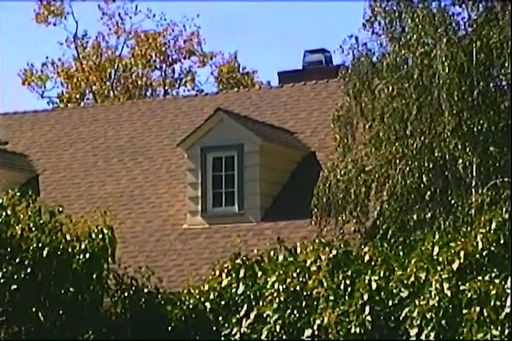 A tan house with dormer windows sits behind dense green hedges under a clear blue sky