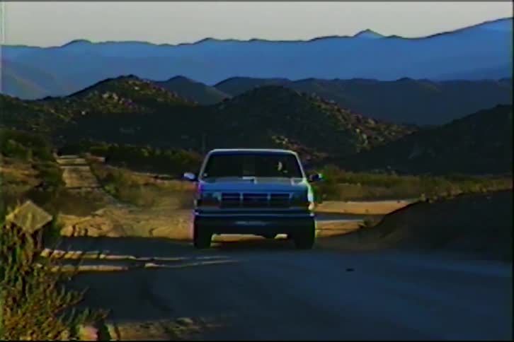 A white ford pickup truck drives along a winding dirt road through a hilly desert landscapethe footage is captured in a vintage vhs style featuring a soft focus and a 43 aspect ratio