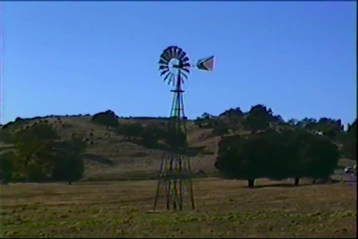 Metal windmill in an open field with hills and trees in the background under a blue sky