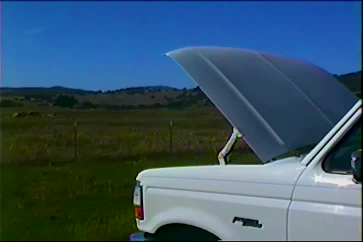 A white truck with an open hood parked on a field outdoors in a rural area during daytime