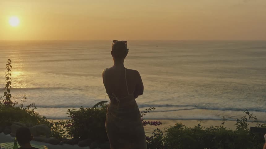 A woman stands on a balcony overlooking the ocean at sunset