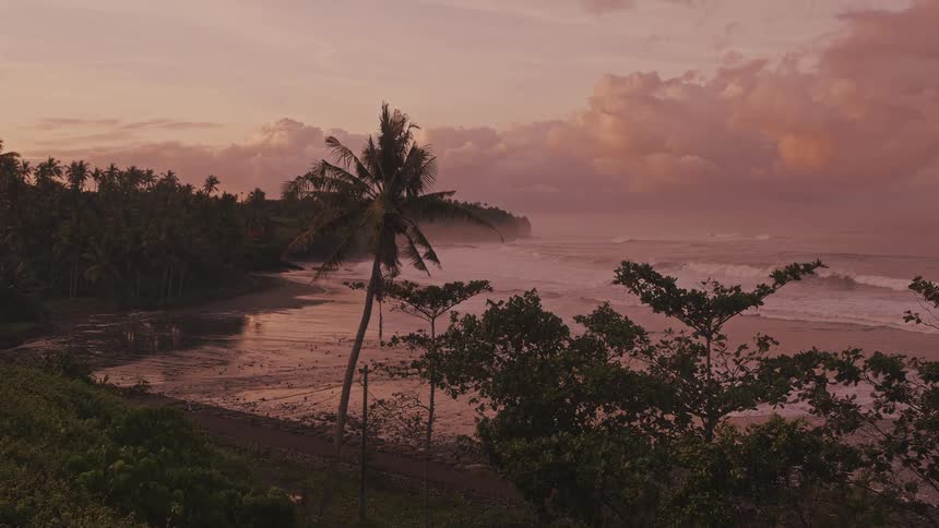 A tropical beach is bathed in a warm pinkishorange glow as the sun sets behind a layer of heavy clouds ocean waves roll gently toward a dark shoreline lined with tall palm trees and lush greenery