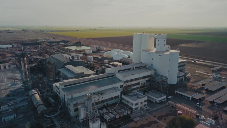 A factory sits along the roadside at midday its series of industrial structures rising behind it as cars drive past in the bright sunlit heat of the day