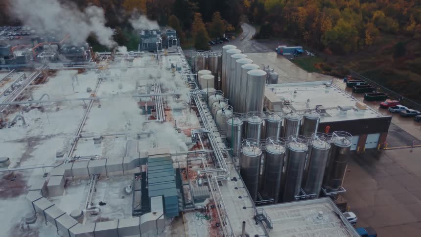 Aerial view of a large industrial factory with multiple silos and rooftop pipes releasing steam