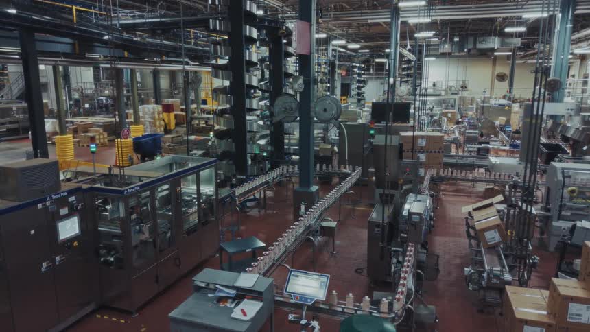 This highangle wide shot shows a bottling production line where rows of plastic containers move along conveyors through automated machinery and packaging stations inside a large industrial manufacturing facility filled with boxes and equipment