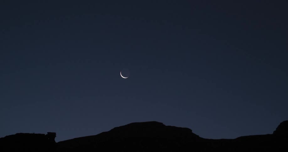 Crescent moon rises over mountains in the night sky as morning arrives