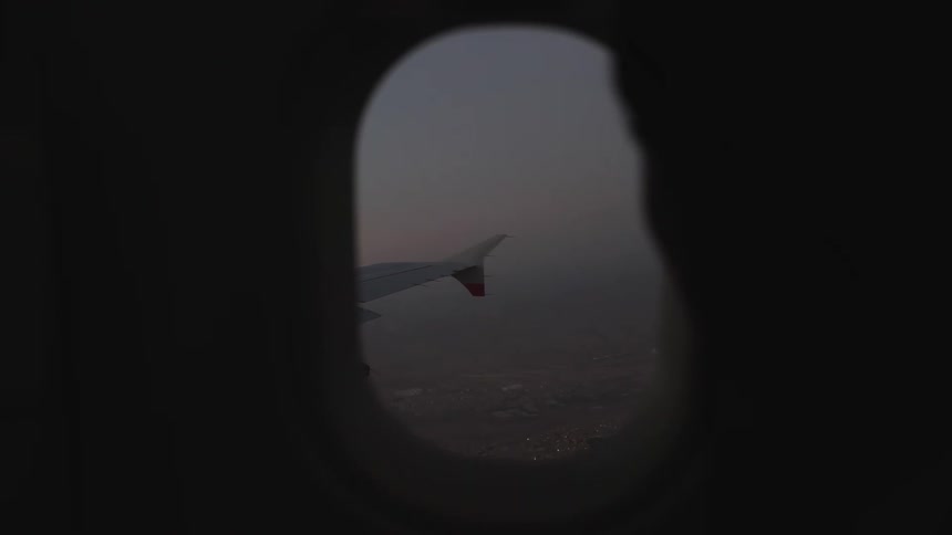 A silhouetted passenger gazes out an airplane window at the wing against a dim hazy sky