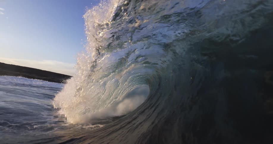 An ocean wave builds and curls into a translucent barrel as it breaks under a clear blue sky the camera moves inside the waves tube before being submerged in a rush of white water and bubbles